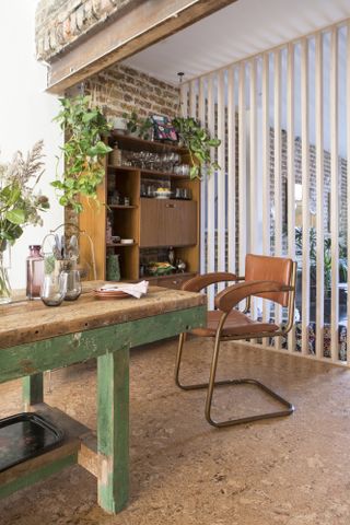 Dining area with cork floor, exposed wood wall, slatted room divider leading to lounge, and old wood table with green painted legs