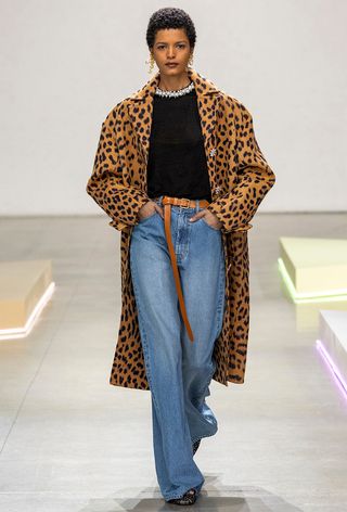 The leopard print coat trend is shown on the fall runway 2024 collection of Zimmerman with a model walking wearing a leopard print coat over a black embellished sweater with jeans, a tan belt, and black shoes