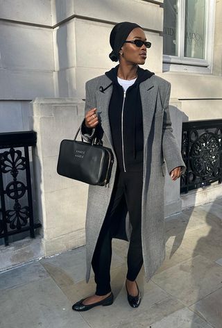 A winter travel outfit is shown in a photo of a woman standing outside wearing a long gray coat over a black zip-up hoodie and white t-shirt styled with black joggers, black ballet flats, a black bowler bag, a black headband, clip-on earrings, a black sunglasses