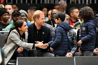 Meghan Markle wearing a gray blazer shaking hands with a boy in a blue coat while sitting next to Prince Harry in the stands at the Invictus Games