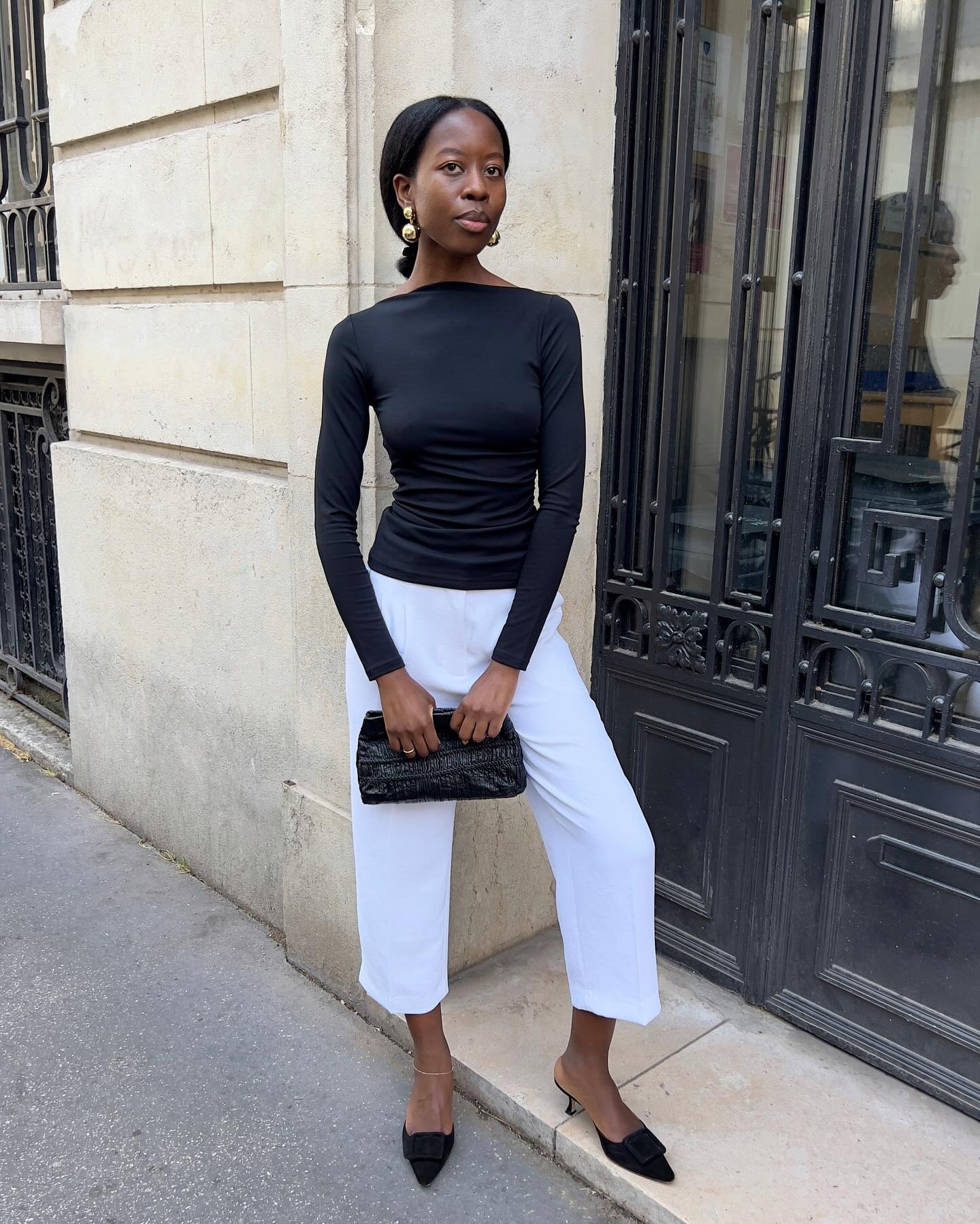 Influencer @sylviemus_ stands outside in Paris wearing a black top with white cropped trousers and black heeled mules. She carries a black bag and wears gold earrings.
