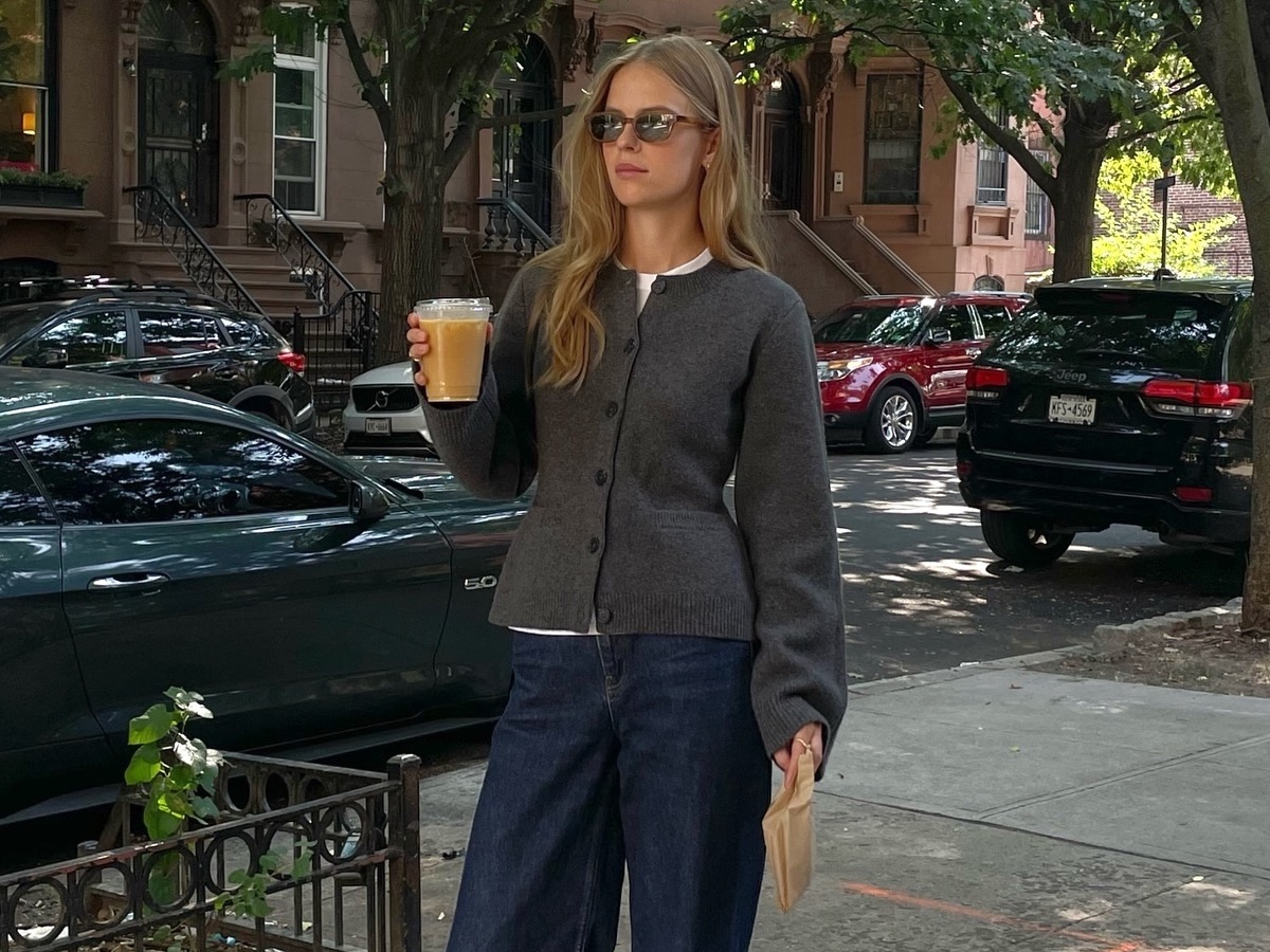 Scandi style influencer and creative Clara Dyrhauge poses on a New York City sidewalk holding a cup of coffee wearing a fitted gray cardigan sweater and relaxed dark-wash jeans