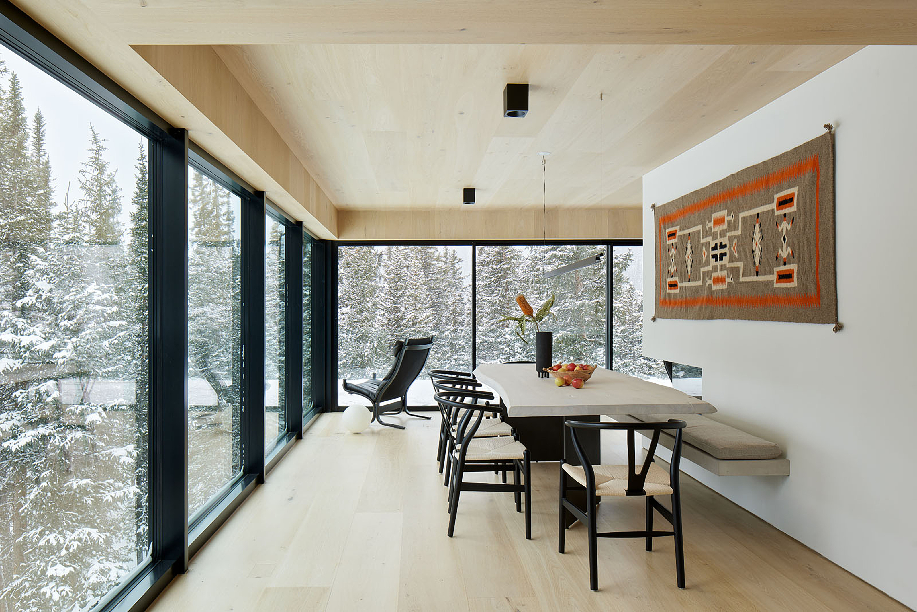 Dining room with white oak ceiling and floor and floor-to-ceiling windows with panoramic views over the mountains, with a simple black and oak table and black chairs