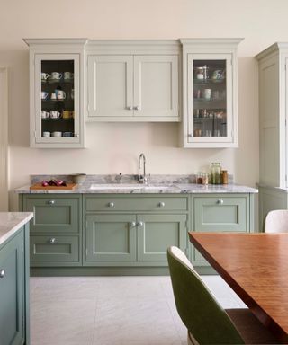 Very pale green eye level kitchen cabinets, with sage green lowers cabinet, marble white and gray counters and wooden table in foreground