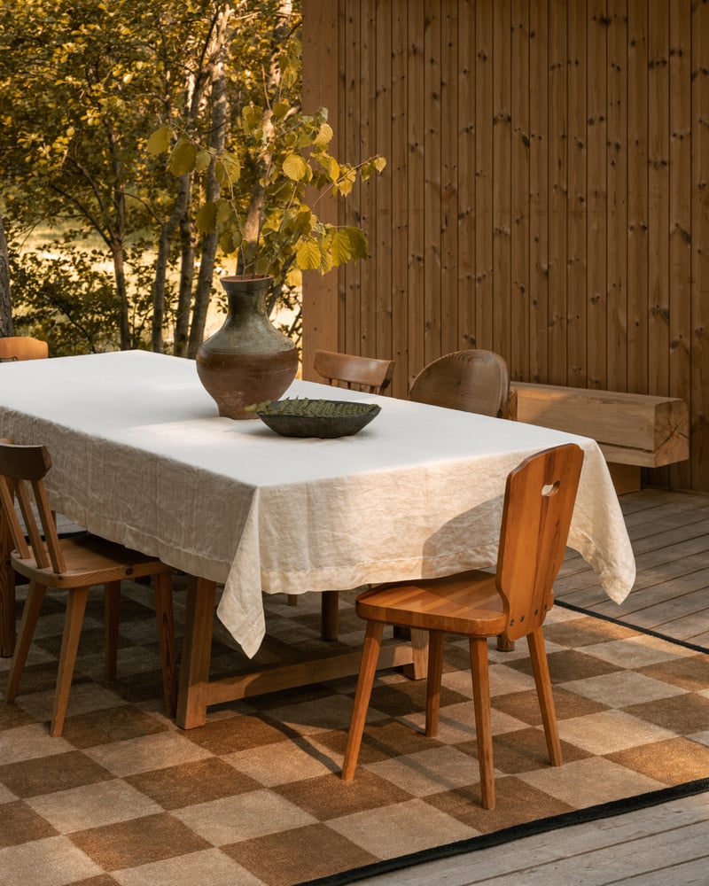 Image of a wooden outdoor patio deck. There is a beige checkerboard jute rug with a wooden dining table and chairs set on it. The table has a white tablecloth and a vase on it.