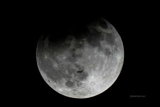 a full moon undergoing a partial lunar eclipse with a shadow in the upper corner of the surface. A plane is visible in the sky flying in front of the moon/