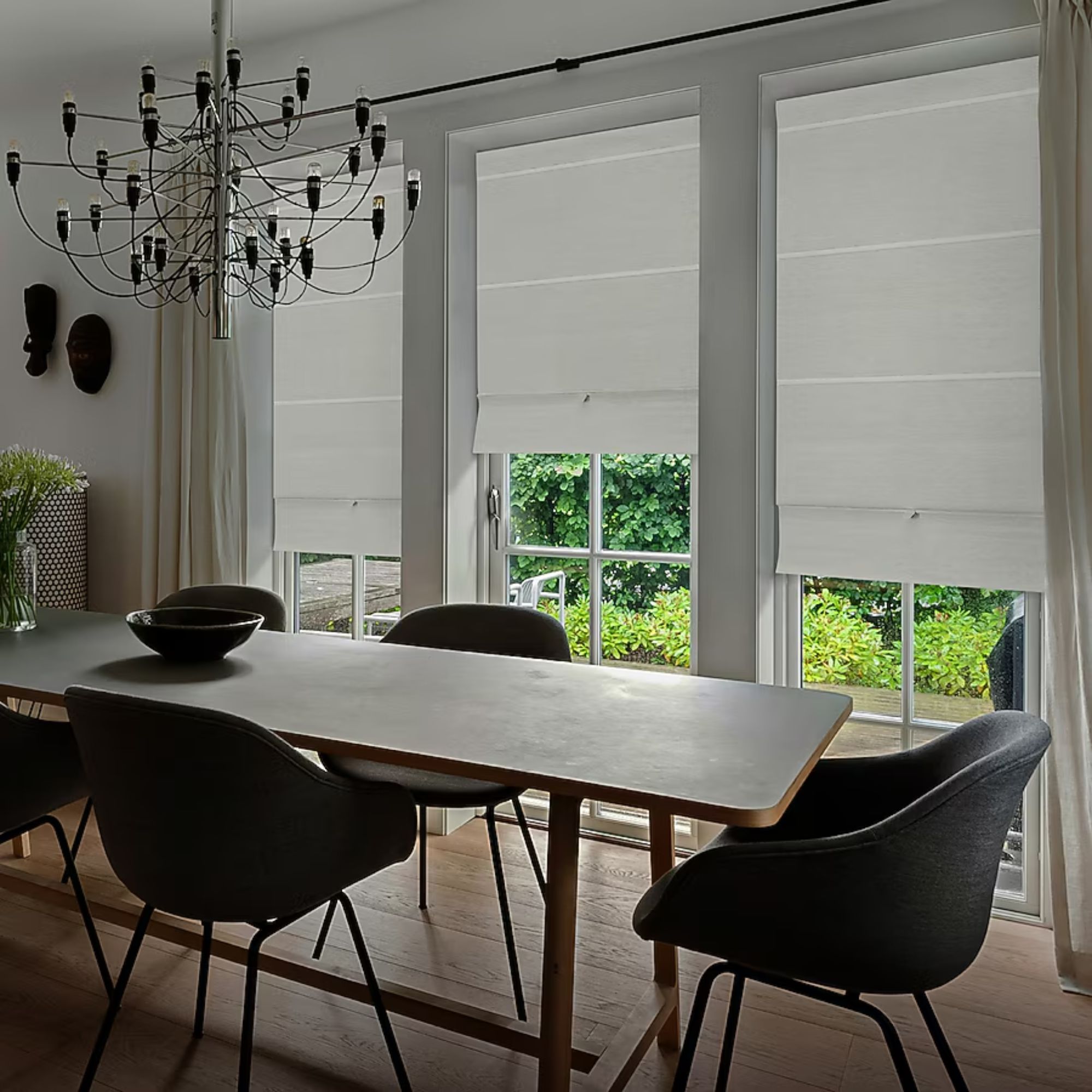 The GoDear Design Blackout Roman Shades shown in white on large windows in a dining space. In the foreground is a large dining table with black dining chairs, flowers, and a large chandelier hanging above.