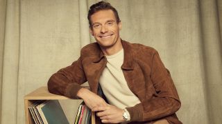Ryan Seacrest leans against a record shelf in American Idol promo. He's wearing a brown suied jacket and white tee. 