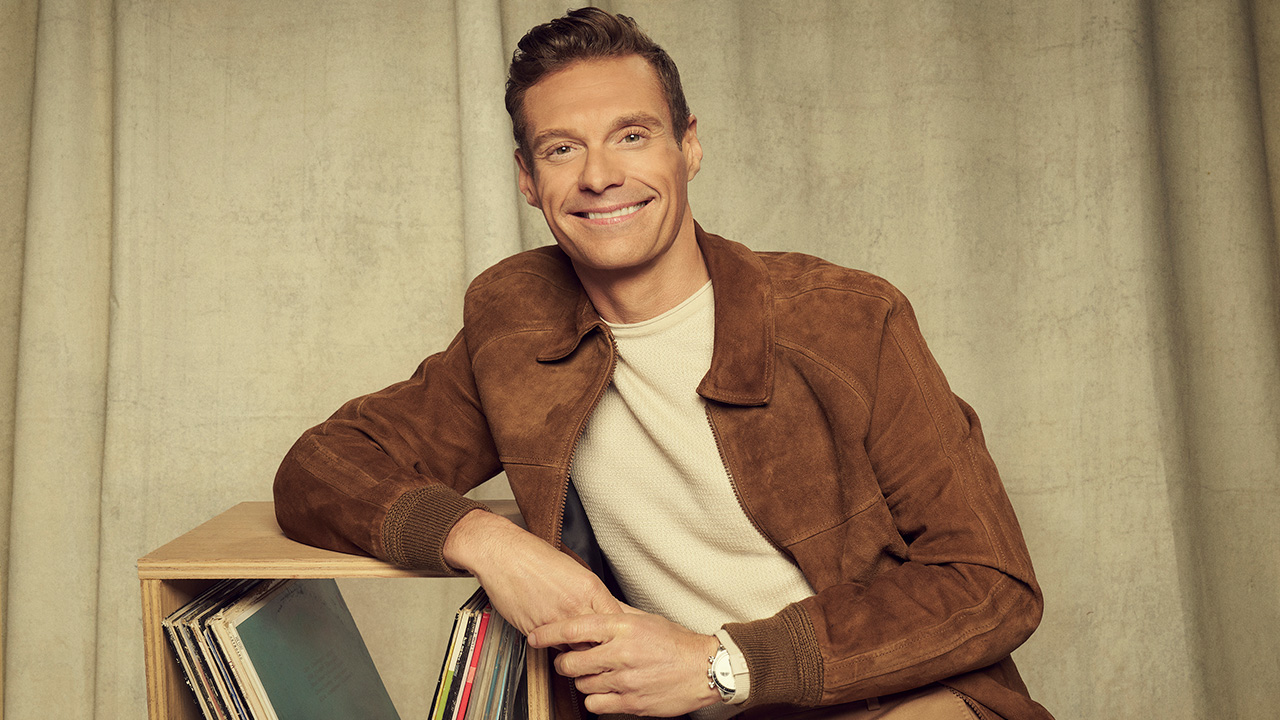 Ryan Seacrest leans against a record shelf in American Idol promo. He's wearing a brown suied jacket and white tee. 