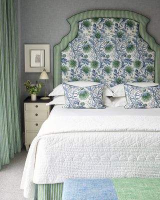 grey bedroom with green and blue patterned fabric, upholstered headboard, green stripe drapes, cream nightstand, white bedding, artwork, cream desklamp