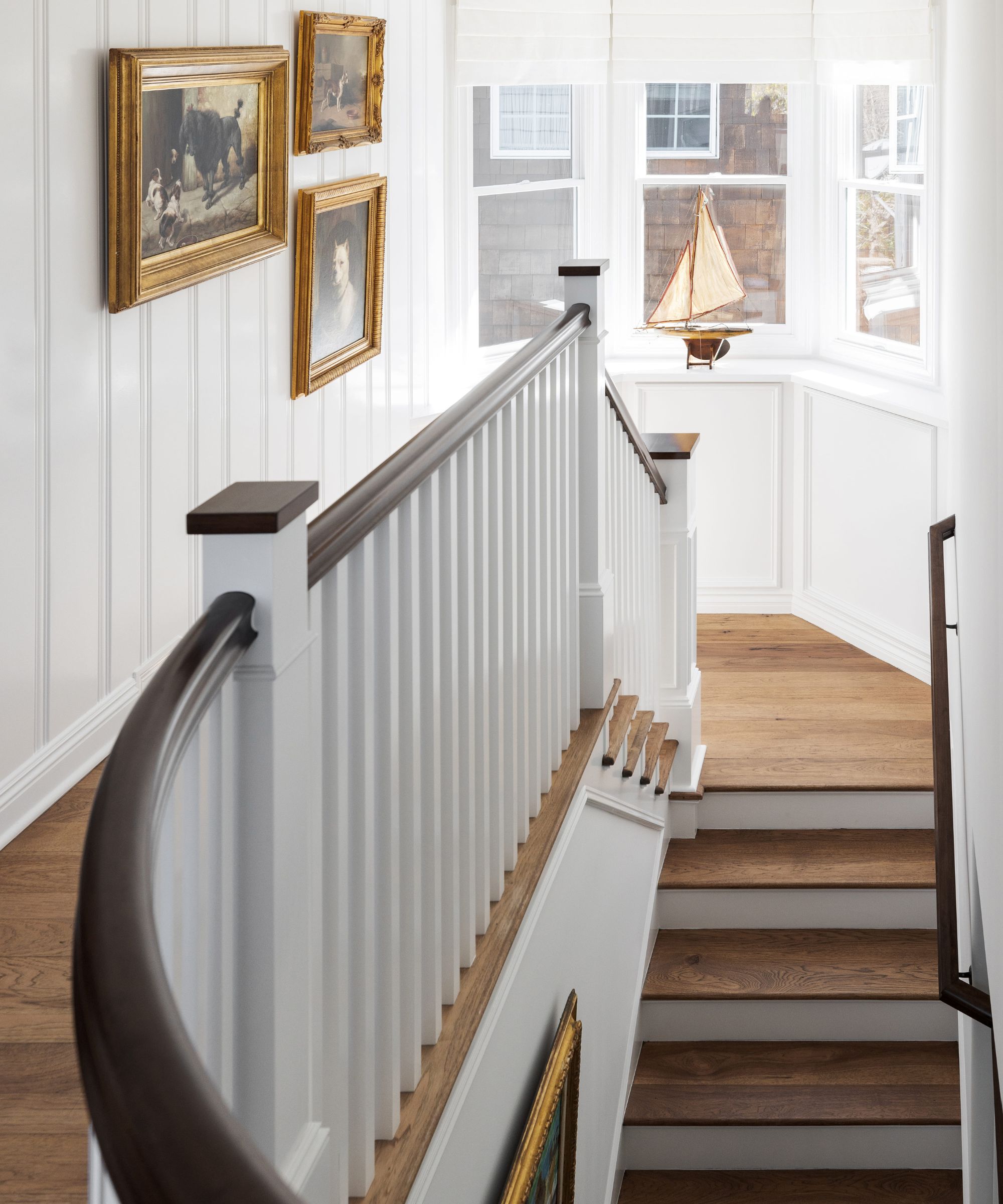 Light-filled staircase with large bay windows, white panelled walls, wooden flooring, vintage artwork on the wall and a vintage ship in the window.
