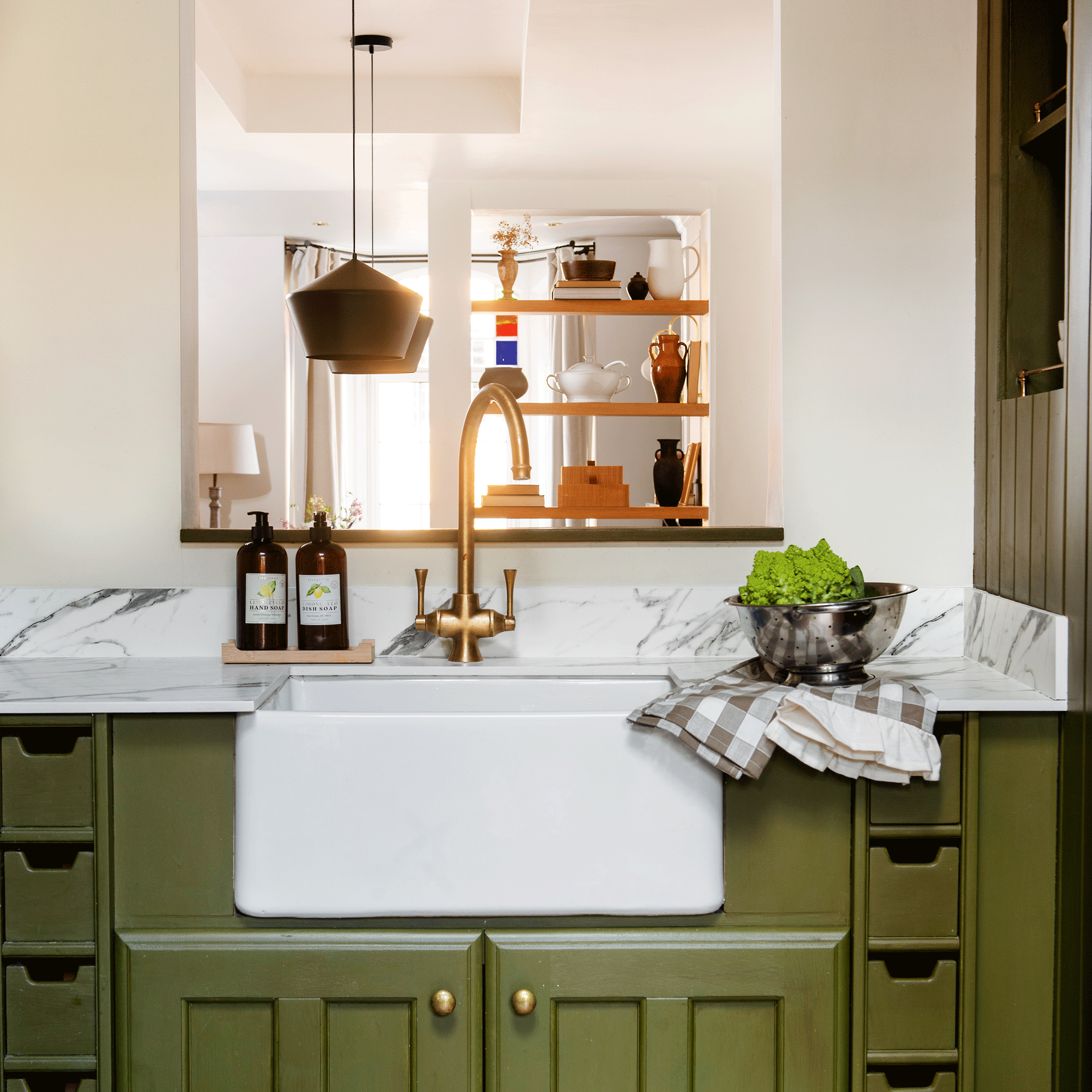 a green kitchen with a Belfast sink and marble countertop with an opening that looks through to the open plan living area