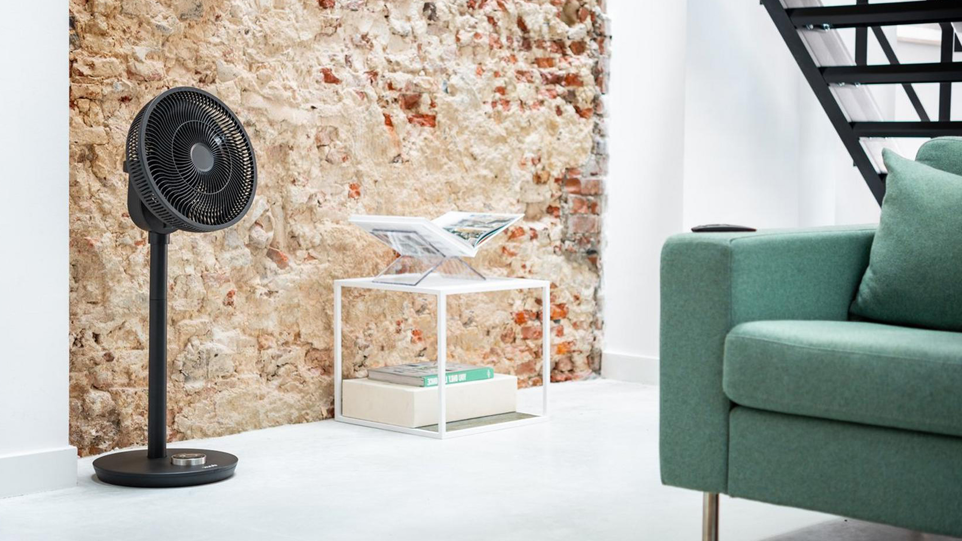 The black Duux Whisper Smart pedestal fan in a room with an exposed brick wall and white floors