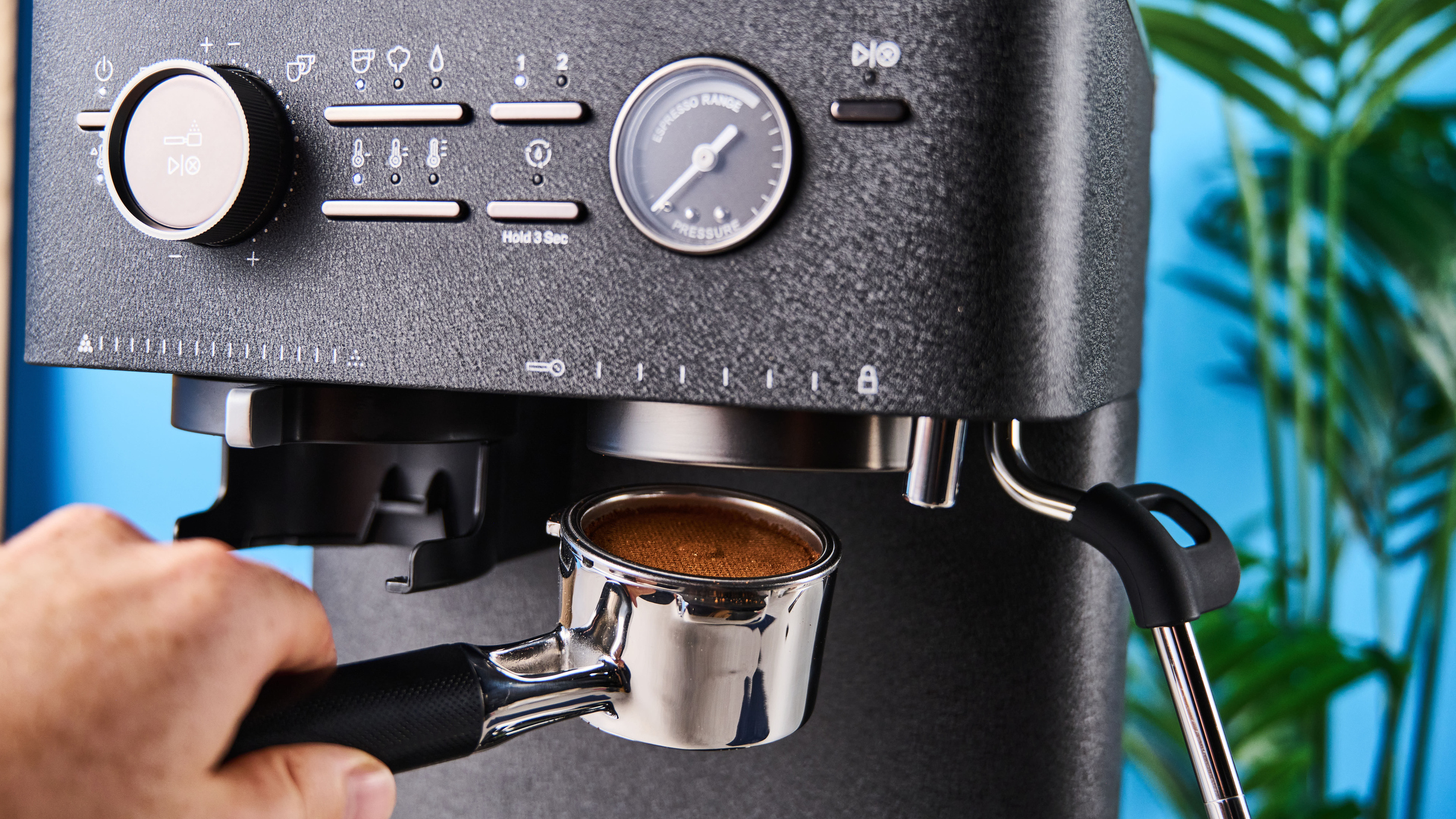 a black espresso machine by kitchenaid photographed against a blue background with silver chrome buttons and a pressure dial and steam wand