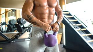 a man with his short off in a gym holding a kettlebell in front of his core muscles which are well defined