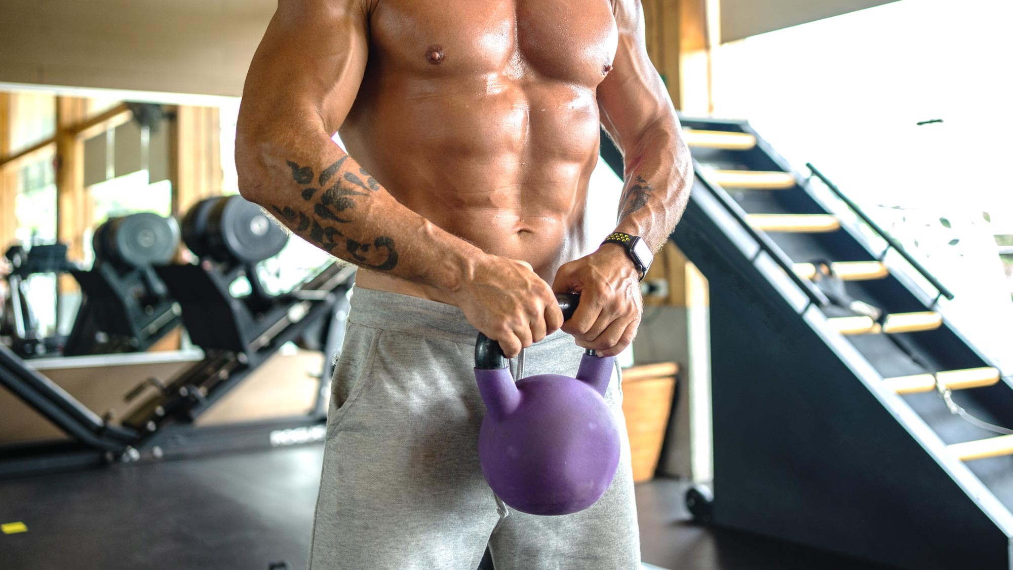 a man with his short off in a gym holding a kettlebell in front of his core muscles which are well defined