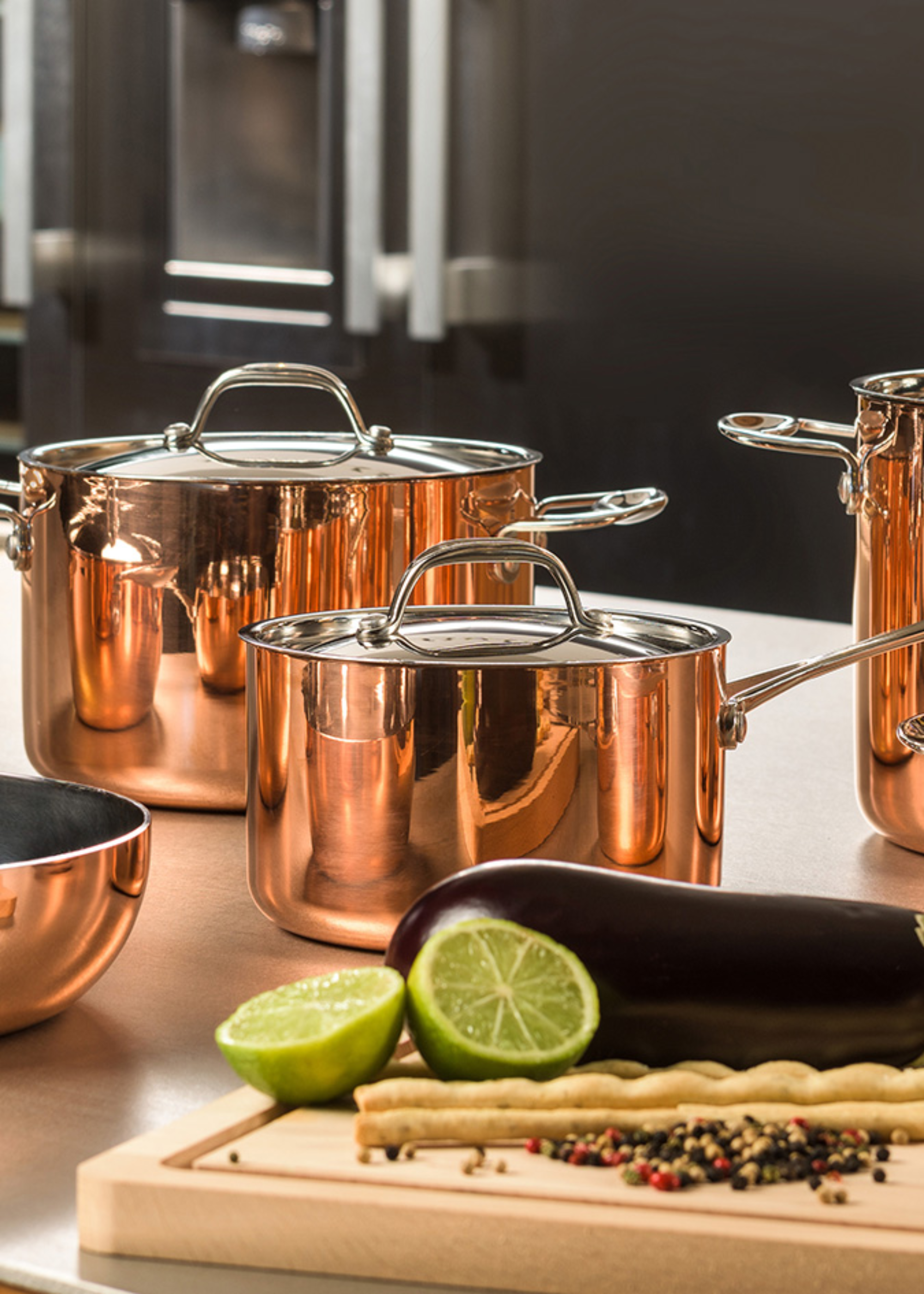 A copper and silver set of pots with lids near a chopping board with spices, limes and an aubergine