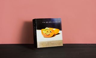 Front cover of the book ' The AOC. Cookbook', black and white design, white and black lettering, orange cut in half, dark wood surface, orange background