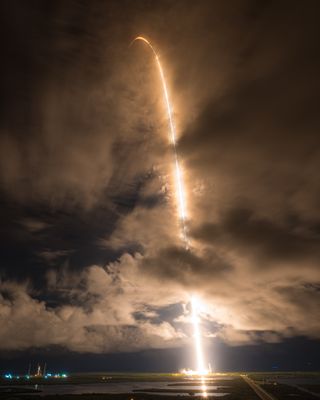 A streak of light shoots in a tall arc from the horizon, rising in the sky, through dimly lit cloud cover.