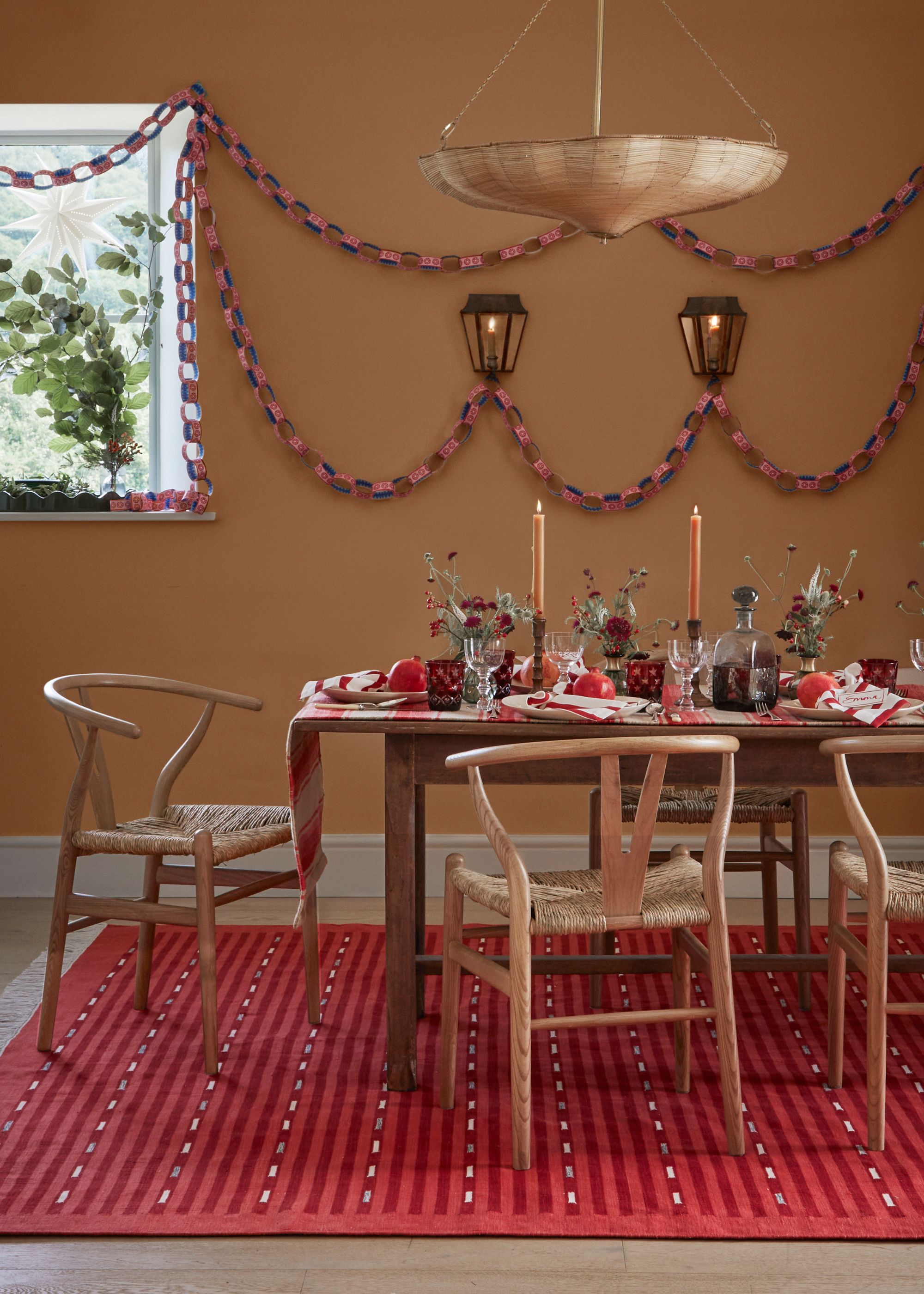 A dining room with light brown walls, a large red rug, and a wooden dining table and chairs with festive decor, including candles and foliage. Paper chain decorations on the wall.