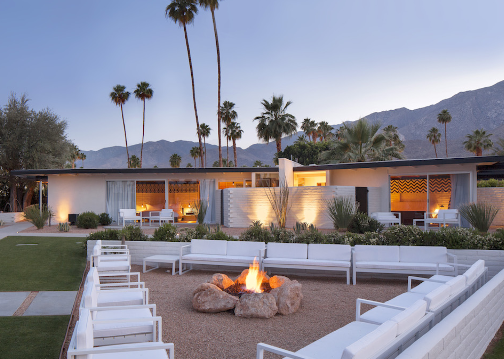 a modern bungalow house surrounded by palm trees with a large outdoor seating area with a rock fire pit in the middle
