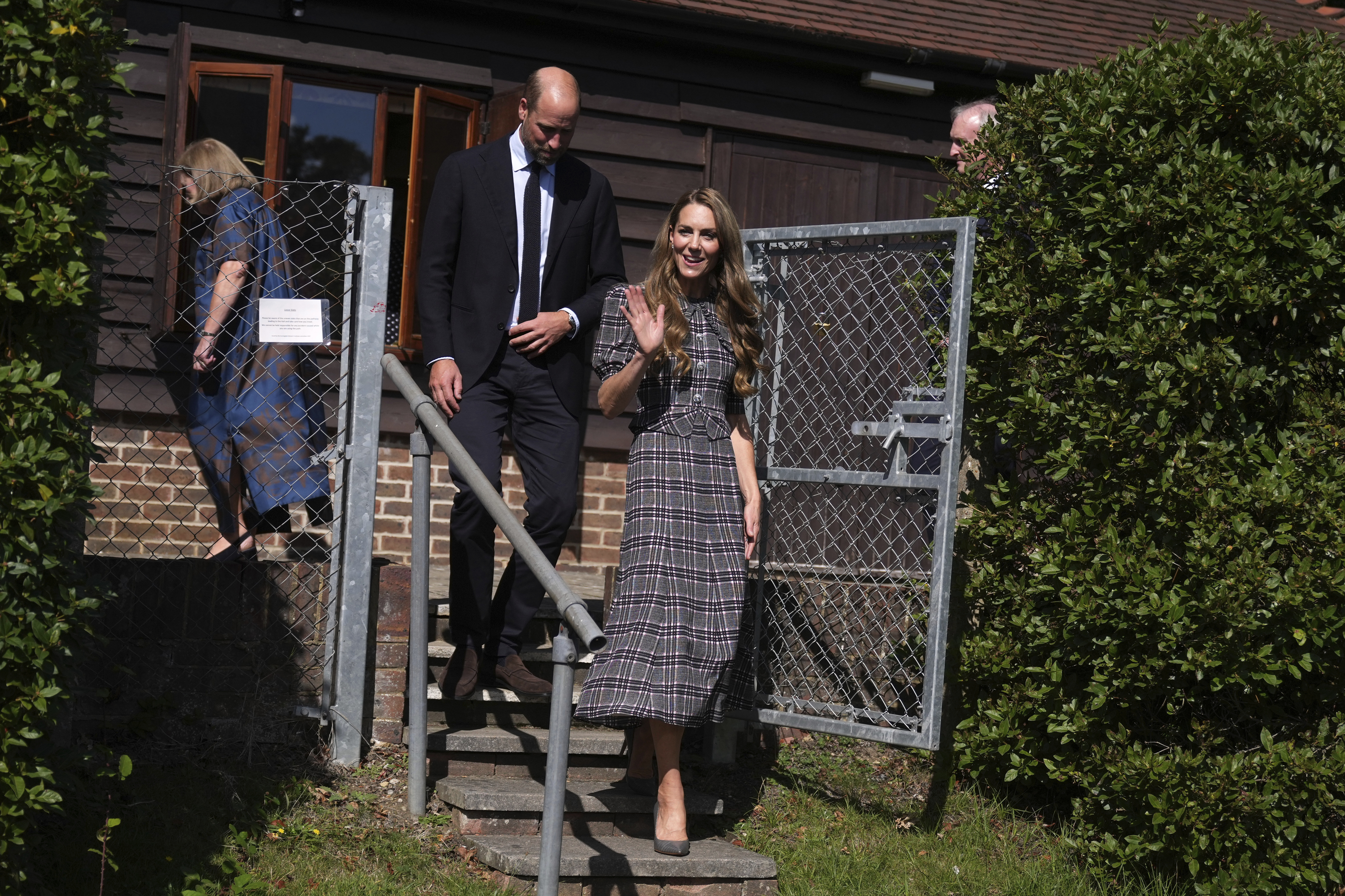 Kate Middleton leaving a brick building wearing a gray plaid dress and waving with Prince William walking down stairs behind her