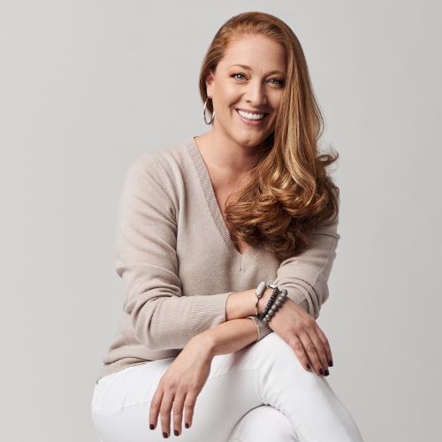 Image of a woman in a beige, V-neck top and white jeans sitting with her legs crossed in front of a gray/beige background.