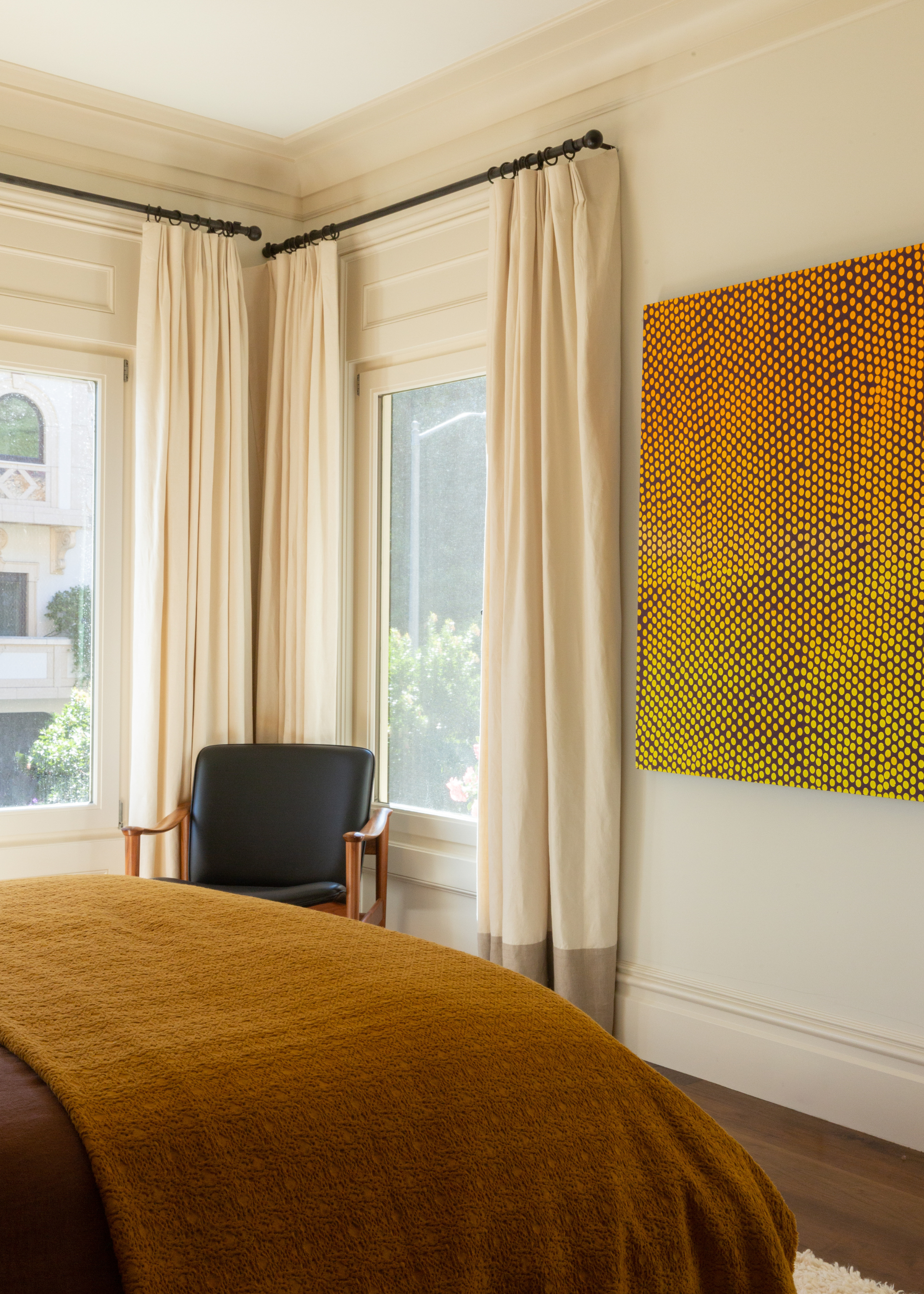 A bedroom with a mustard duvet, a side chair, white curtains, and a vibrant painting