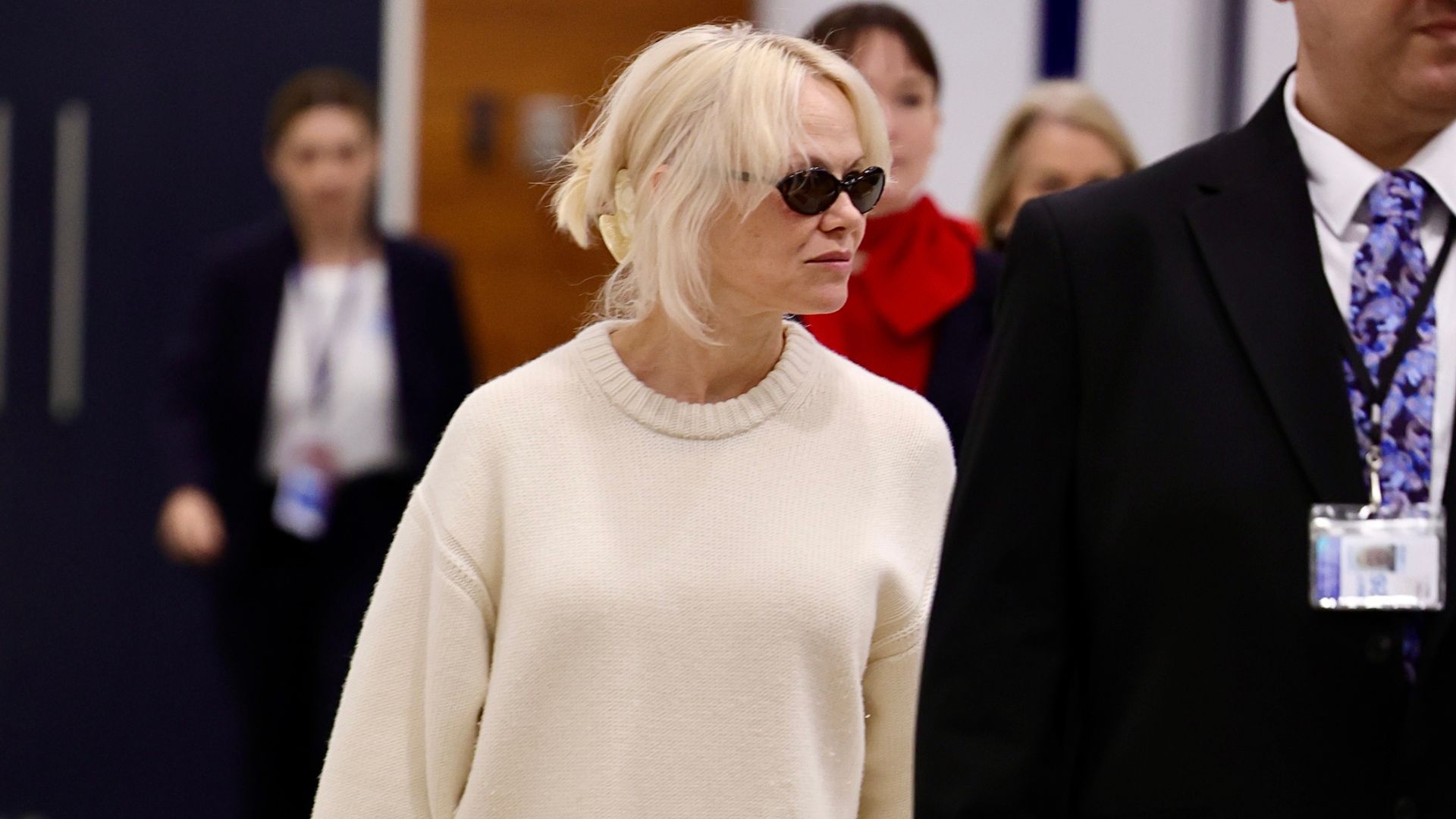 Pamela Anderson walks through the airport wearing white trainers with a wicker bag, a white jumper and white trousers.