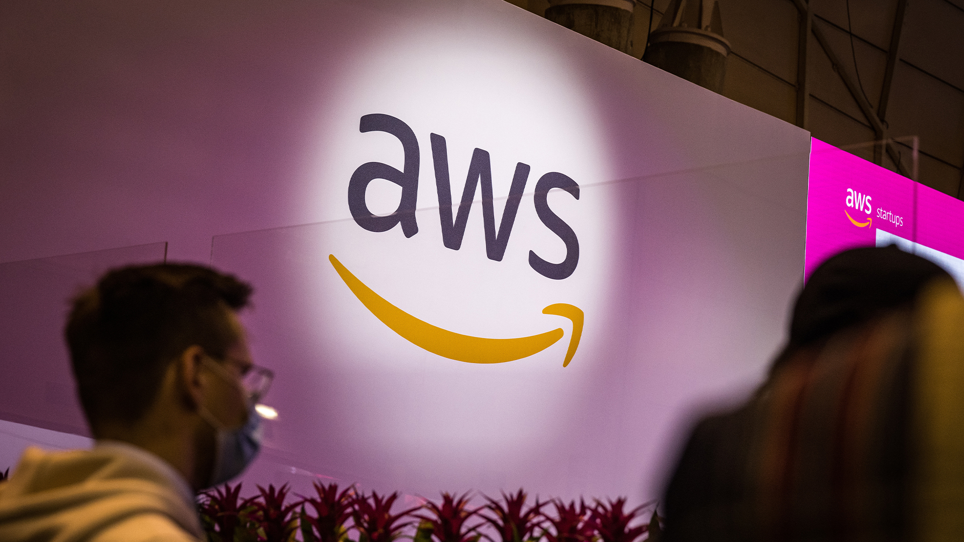 Amazon Web Services (AWS) logo pictured on a wall at the Lisbon Web Summit with attendees walking by.