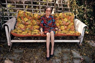 View of Devoe Money wearing a multicoloured dress, glasses and black shoes sitting on a floral sofa bed outside during the day