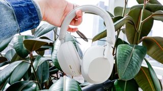 Sony XM5 headphones in ecru being held in mid air with green plant in background