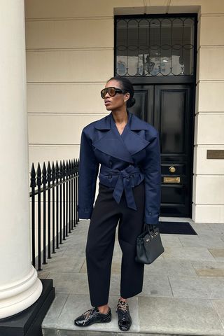 Spring trench coats shown in a photo of a woman standing outside in London in on a stoop wearing aviator sunglasses, a navy cropped trench coat, black barrel-leg pants, a black handbag, and black sneakers