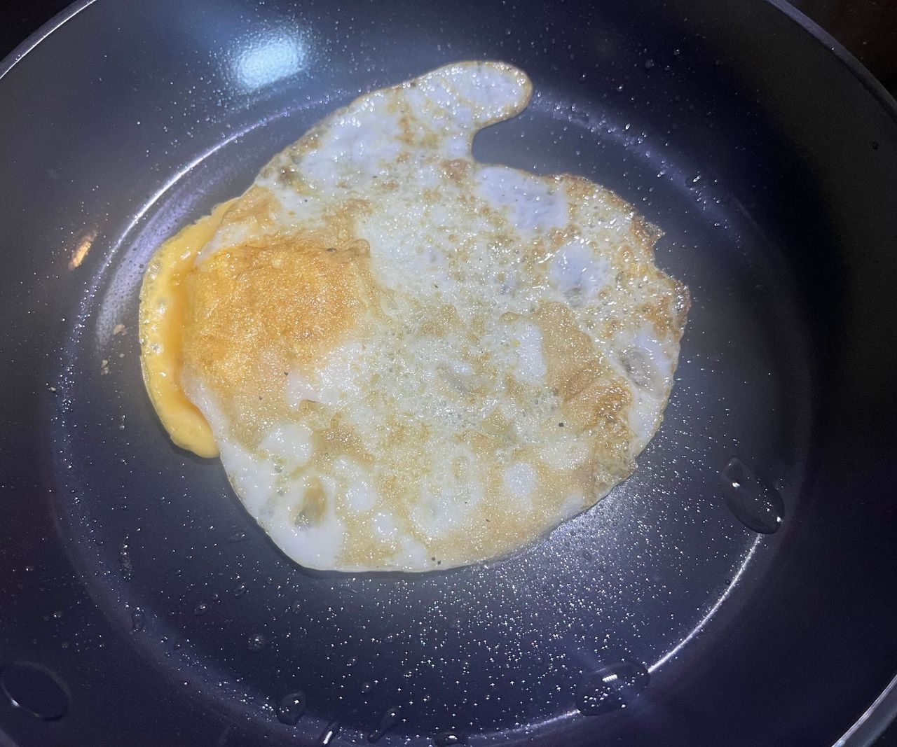 A black ceramic frying pan on a black stovetop with an upside-down fried egg with a golden base.