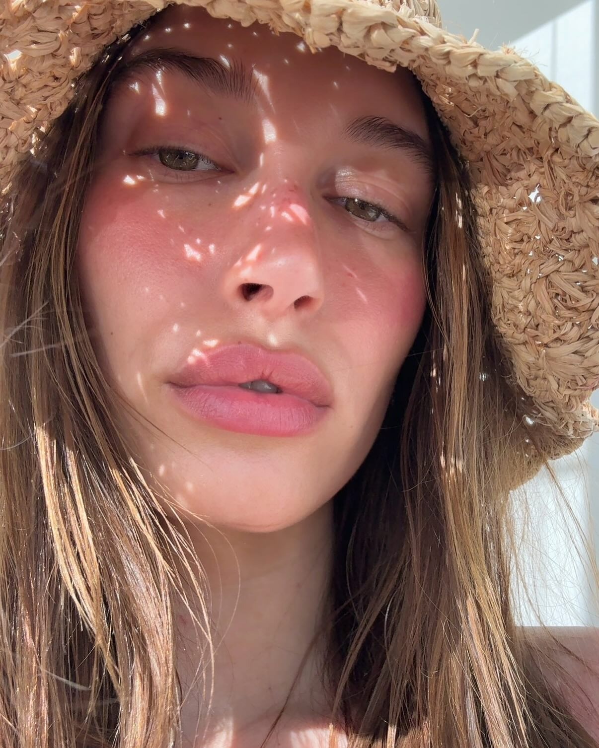 Hailey Bieber wearing a straw hat with sunburnt-style pink blusher on her cheeks and nose