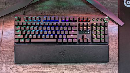 A mechanical keyboard with RGB backlit keys displaying various colors on a wooden desk. It features a wrist rest and a sleek, modern design.