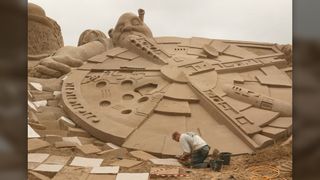 A man sculpts sand into an enormous Millennium Falcon with Shrek in the background.