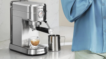 The Casabrews CM5418 on a modern white counter next to a steel milk pitcher and a shot of espresso