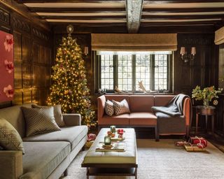 A cosy living room with two sofas, with exposed ceiling beams, a tall decorated Christmas tree and fireplace with a lit fire.