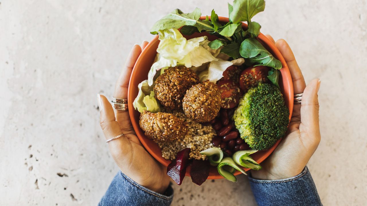 Woman&#039;s hands holding a healthy bowl of food, which includes falafel, salad and broccoli