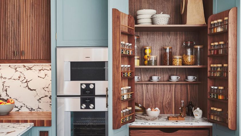 This image features a compact pantry integrated into a modern kitchen design. The pantry doors, crafted from rich wood with vertical grooves, open to reveal an organised storage space. The interior showcases wooden shelving stocked with glass jars containing spices, grains, and preserved goods, alongside neatly arranged cups and bowls.