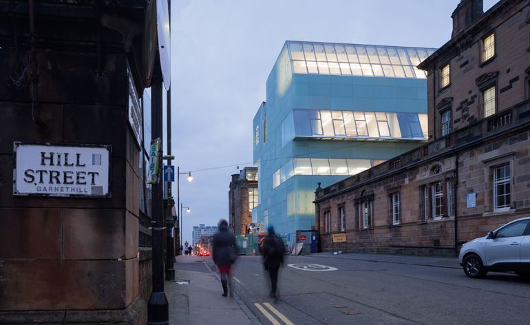 Photograph captured from the corner of hill street with blurred image of 2 people walking on the street and on the right 3 buildings with the Reid building in the center.