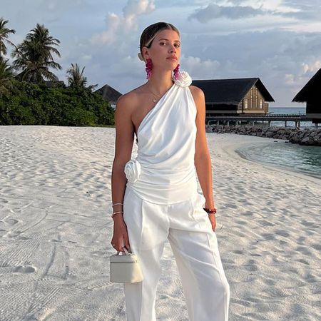 Sofia Richie Grainge wearing a white one-shoulder top, white trousers, and an off-white Loro Piana Extra Pocket top-handle vanity case bag on her honeymoon.