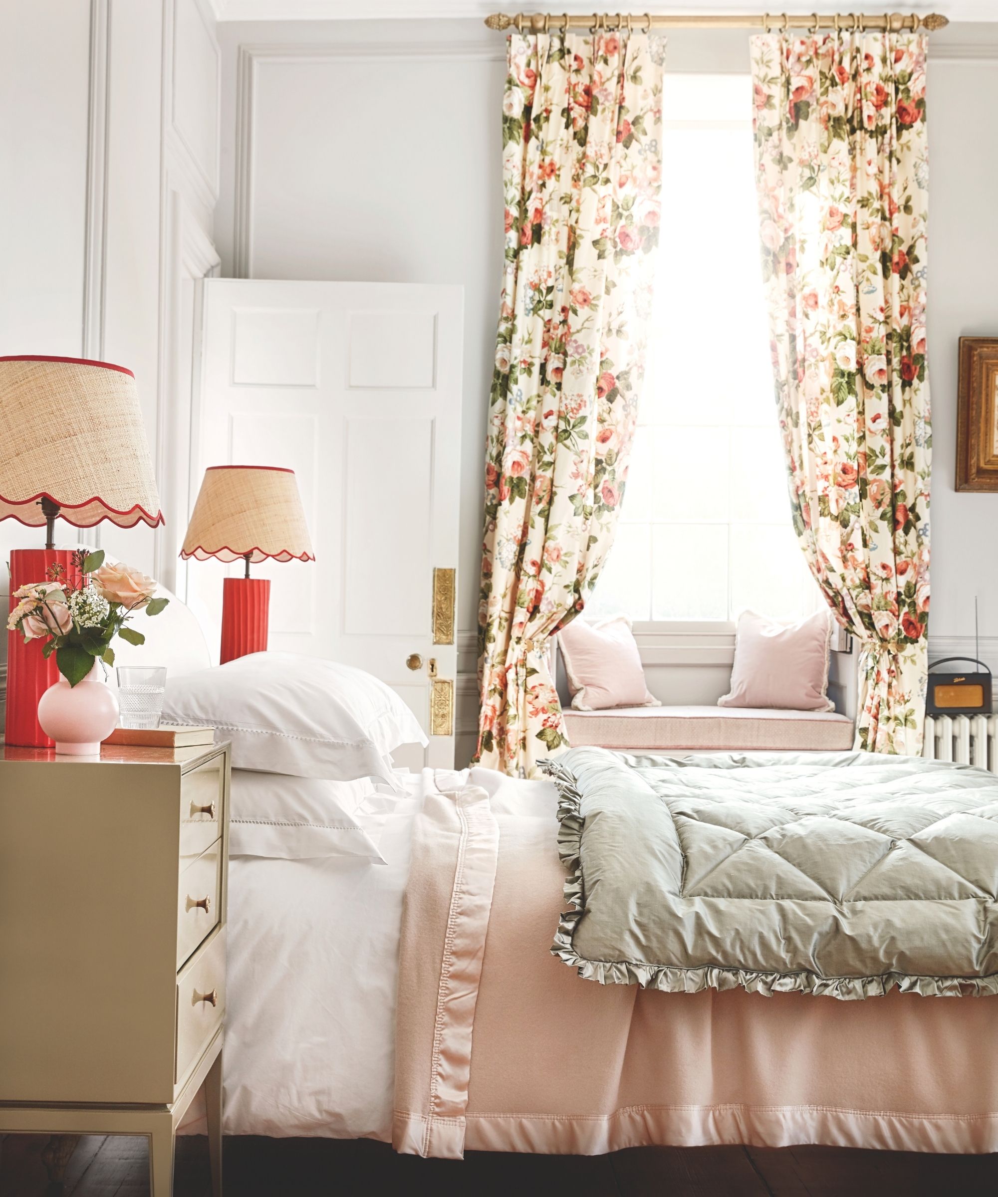 Bedroom with white walls, pale pink quilt, floral curtains, and matching scalloped lamps