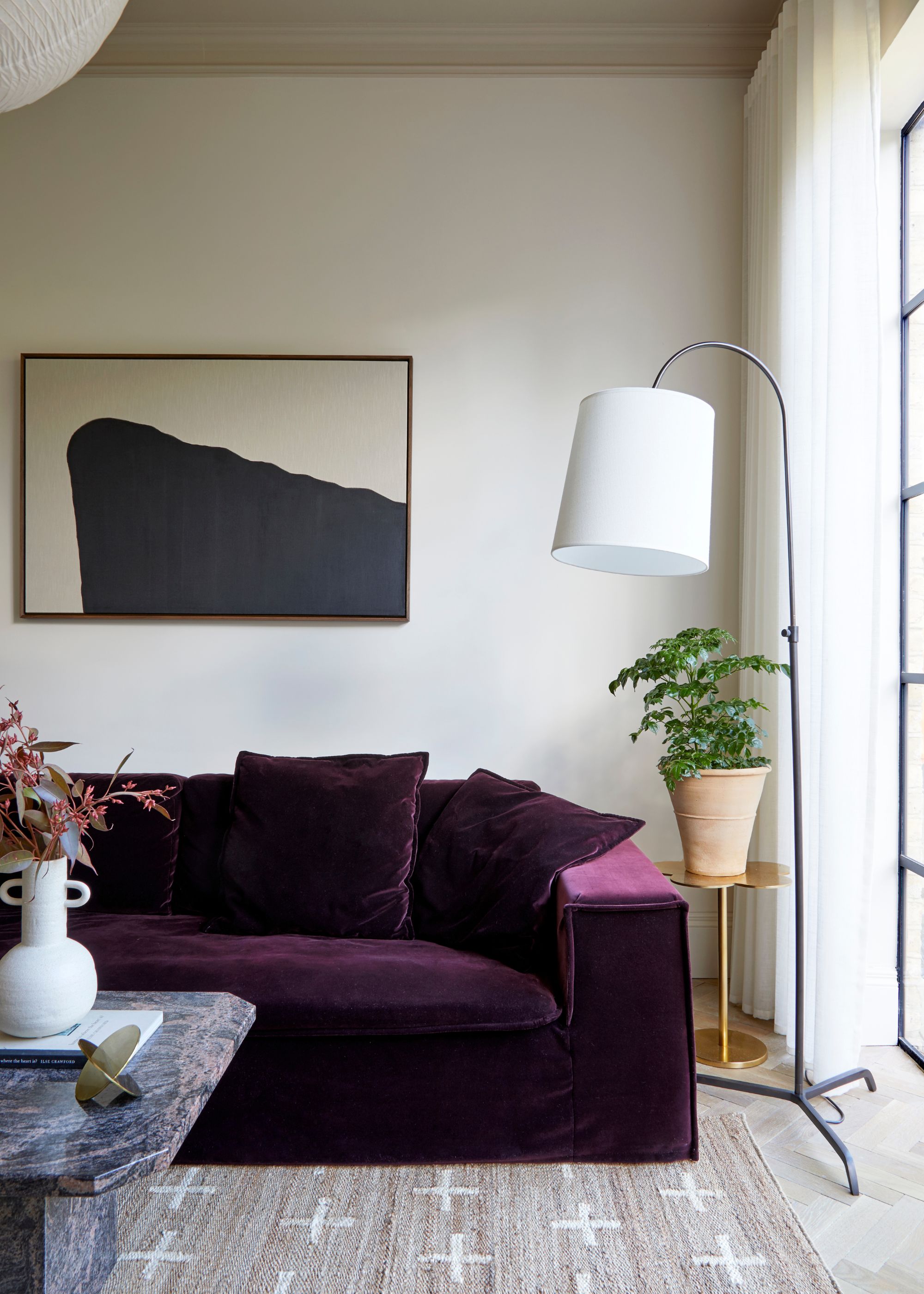 A modern living room with soft neutral walls, a dark plum velvet sofa, a jute rug, a minimalist floor lamp, and black and beige abstract artwork on the wall.