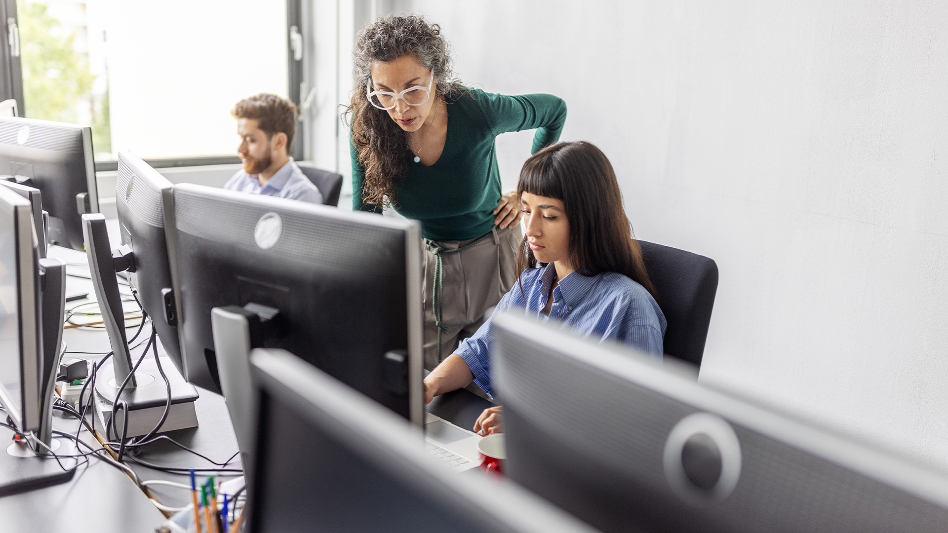 IT service management (ITSM) professionals working on computers in an office space, with female worker looking over colleague's shoulder at desktop monitor.