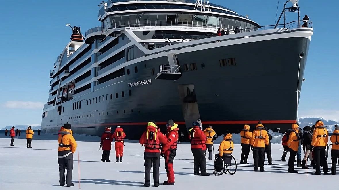 dozens of people in red and orange thermal coats stand on ice around a large black cruise ship