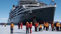 dozens of people in red and orange thermal coats stand on ice around a large black cruise ship