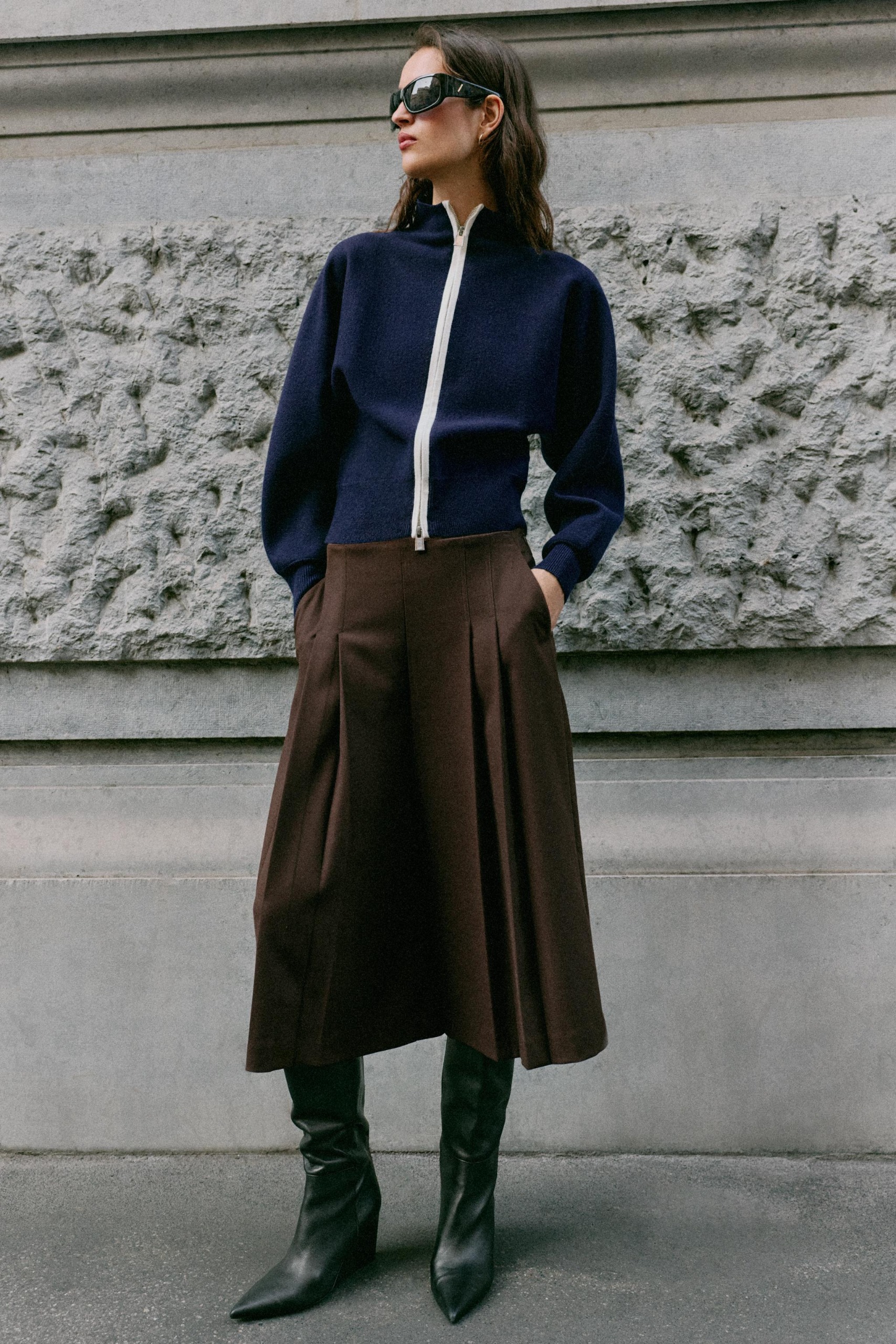 Model wears pleated brown culottes, navy jacket, and tall black boots.