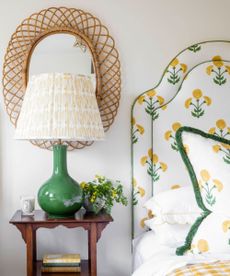 A bright bedroom with white walls, with a white bed headboard with green and yellow flowers and matching pillow, wooden bedside table and green lamp with white lampshade and rattan oval mirror